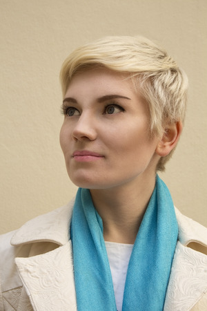Close-up stylish portrait of beautiful young woman blonde wearing blue scarf and white coat isolated on beige background in profile. Girl with short hair. Haircut. Hairstyle. Fringe. Professional nude makeup. Fashion fresh make-up. Model shooting.の写真素材