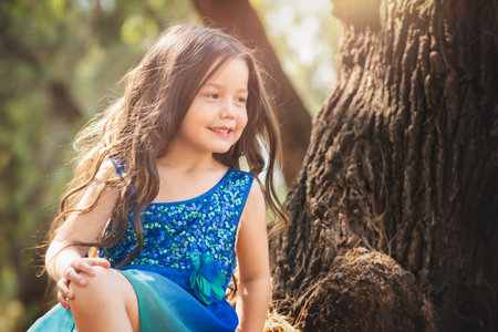 beautiful happy girl playing in the park with blue dress without shoes sitting on tree laughing screaming with happiness in family enjoying children's dayの写真素材