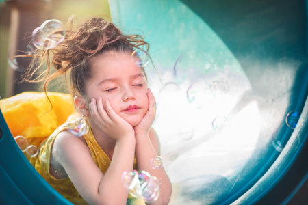 beautiful junior little girl playing in the park in yellow dress laughing with bubbles, screaming with happiness in family enjoying children's dayの写真素材
