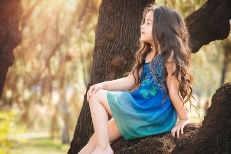 beautiful happy girl playing in the park with blue dress without shoes sitting on tree laughing screaming with happiness in family enjoying children's dayの写真素材
