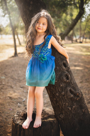 beautiful happy girl playing in the park with blue dress without shoes sitting on tree laughing screaming with happiness in family enjoying children's dayの写真素材