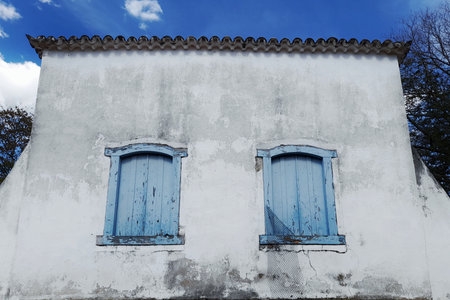 Facade of an old building from the 18th century in white color with dark spots and two blue windows, on the sides of the building there is some vegetation and the blue sky aboveの写真素材
