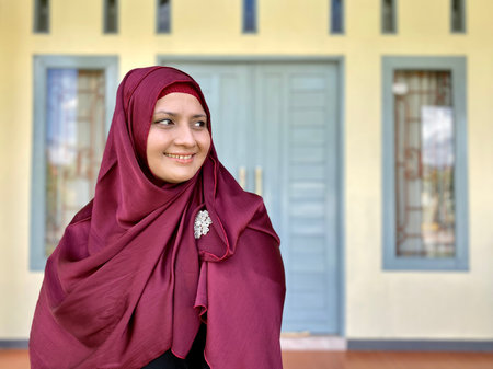 Portrait young woman wearing red hijab, looking away and smiling in front of houseの写真素材