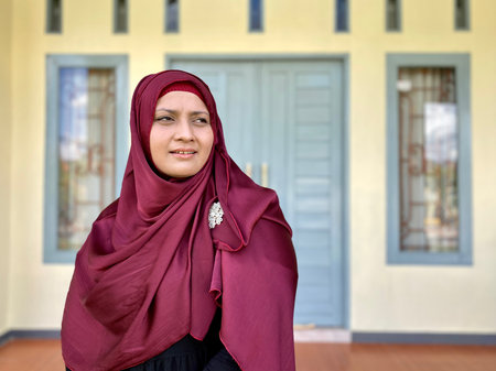 Portrait young woman wearing red hijab, looking away and smiling in front of houseの写真素材