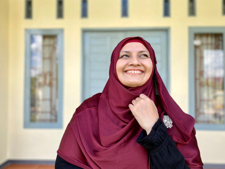 Portrait young woman wearing red hijab, looking away and smiling in front of houseの写真素材