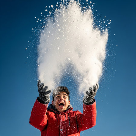 Young man in red jacket throwing snow at camera on blue sky backgroundの素材