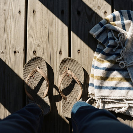 Worn flip-flops and a faded towel resting on a dusty, sun-drenched wooden deck.の素材