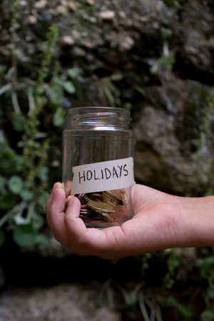 A hand with a glass jar with coins inside it labeled "Holidays" with a stone background and plants.の写真素材