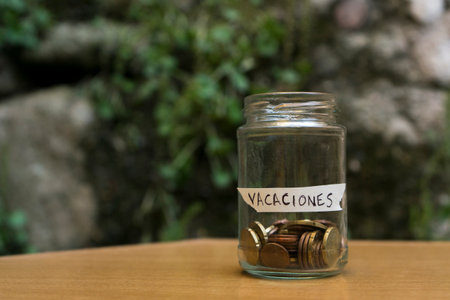 A glass jar with coins inside labeled "Vacations" (traduction "Holidays") with a background of wood, stone and plants.の写真素材