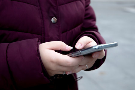 Young girl with a mobile device in her hands typing, wears maroon winter clothes while on the street.の写真素材