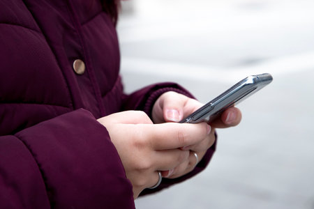 Young girl with a mobile device in her hands typing, wears maroon winter clothes while on the street.の写真素材