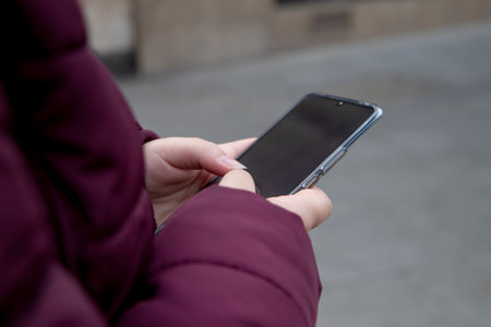 Young girl with a mobile device in her hands typing, wears maroon winter clothes while on the street.の写真素材