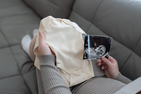 Pregnant woman with ultrasound images and baby bodysuit on sofa. Waiting and preparing for baby...