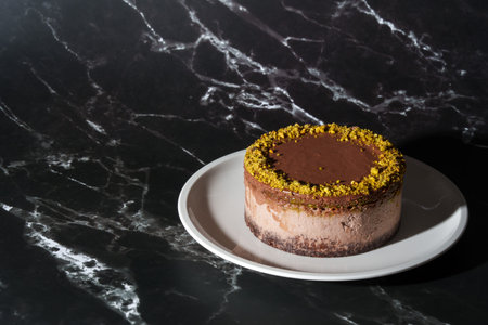 Chocolate cheesecake with pistachio paste and kataifi dough filling covered in chocolate ganache on white plate. Black background with copy space.の写真素材