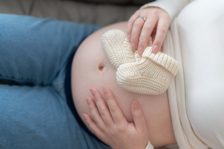 Pregnant woman holding small knitted baby shoes on her belly relaxing at home on couch. Concept of anticipation, motherhood, and preparing for a new life.の写真素材