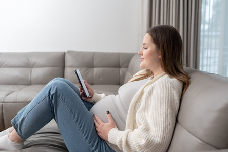 Pregnant woman sitting on sofa at home using smartphone for online shopping and digital services with copy space. Concept of maternity, digital connection, technology and modern ecommerce convenience.の写真素材