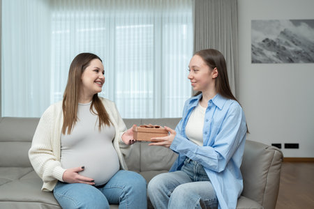 Young woman giving a gift to her pregnant friend while sitting on a couch and smiling in a cozy home. Concept of maternity, celebration, friendship, anticipation, and emotional connection.の写真素材