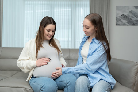 Smiling pregnant woman in third trimester sitting on sofa while her female friend touches her belly with care. Concept of maternity, friendship, connection, support and emotional bonding.の写真素材