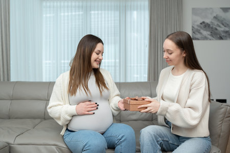 Young woman giving a gift to expecting mother while sitting on a couch and smiling in a cozy home. Concept of pregnancy, maternity, celebration, friendship, anticipation, and emotional connection.の写真素材