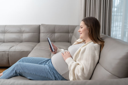 Pregnant woman reclining on a couch, using a smartphone for online call with doctor, enjoying a moment of digital convenience. Concept of technology, maternity and digital healthcare servicesの写真素材