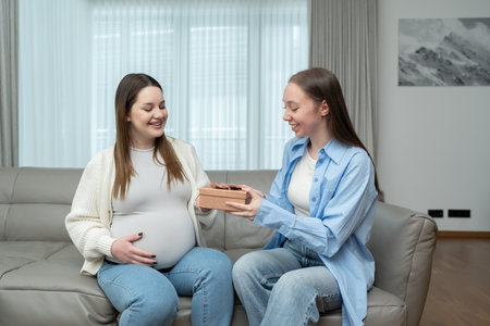Smiling pregnant woman receiving a gift from her female friend while sitting on a couch in a cozy home. Concept of maternity, celebration, friendship, anticipation, and emotional connection.の写真素材