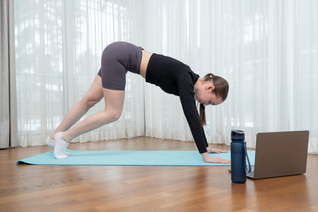 Young woman practicing yoga indoors with online guidance using laptop. Woman doing stretching...