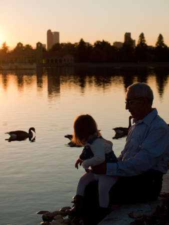 Girl toddler with grandfather by lake at sunset.の写真素材