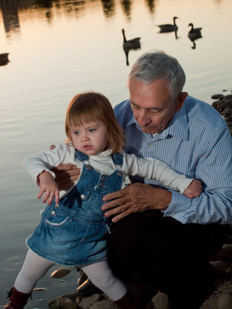 Girl toddler with grandfather by lake at sunset.の写真素材