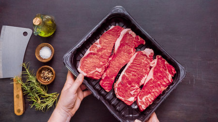 Raw New York strip steaks in black plastic meat packaging container.