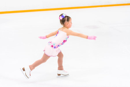 Little girl in a pretty pink dress with flowers practicing figure skating moves on an indoor ice rink.の写真素材