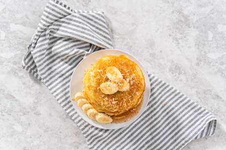 Stack of freshly baked coconut banana pancakes garnished with sliced bananas, toasted coconut, and maple syrup.の写真素材