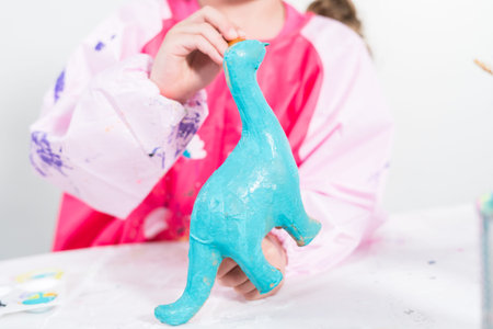 Homeschooling during   lockdown. Little girl painting paper mache dinosaur figurine with acrylic...