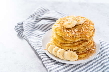 Stack of freshly baked coconut banana pancakes garnished with sliced bananas, toasted coconut, and maple syrup.の写真素材