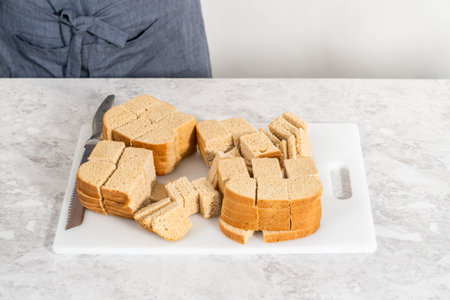 Cutting stale bread with a bread knife on a white cutting board to make bread pudding.