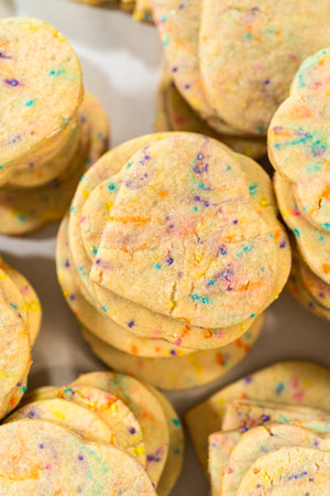Large quantity of sprinkle-filled sugar cookies are neatly stacked on a baking tray, awaiting their packaging into the rustic brown paper boxes.の写真素材