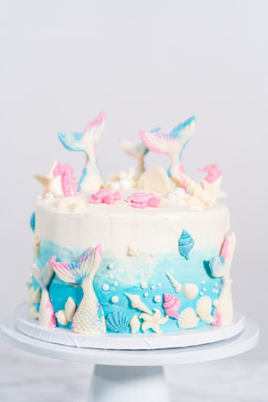 Mermaid-themed 3 layer vanilla cake decorated with chocolate mermaid tails and seashells on a white cake stand.の写真素材