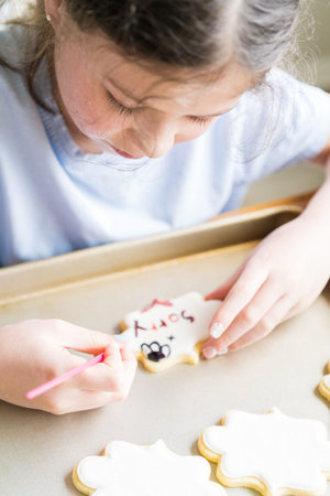 A heartwarming scene of a little girl carefully writing Sorry on sugar cookies with food coloring,...