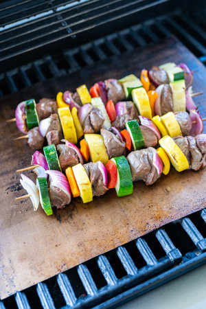Skewered beef and fresh veggies sizzle on a copper grill mat over a gas outdoor grill.の写真素材