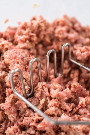 Mixing ingredients with a potatoe masher in a glass mixing bowl to prepare oven-baked meatballs.