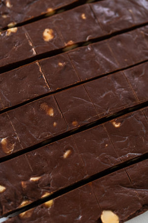 Cutting chocolate macadamia fudge with a large kitchen knife into square pieces on a white cutting...