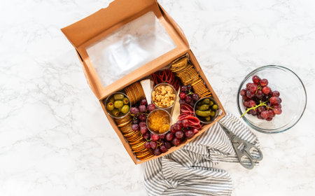Flat lay. In the process of assembling, a woman expertly curates this charcuterie box, showcasing...