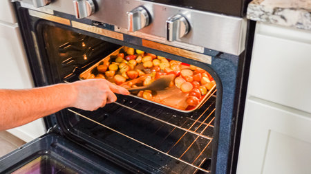 Within the setting of a modern kitchen, a young man is immersed in preparing dinner, currently roasting seasoned rainbow potatoes in the oven, an important step towards a delightful meal.の写真素材