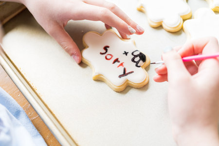 A heartwarming scene of a little girl carefully writing Sorry on sugar cookies with food coloring, the cookies beautifully flooded with white royal icing.の写真素材