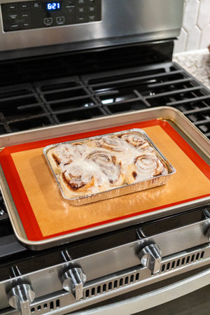 Just out of the oven, cinnamon rolls cool on a silicone baking mat, their icing glistening under the...