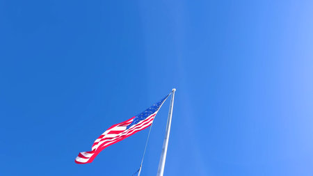 A striking image of the American flag soaring in the wind, set against a vivid blue sky with ample...