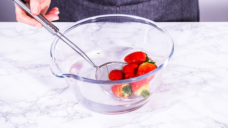 Ripe strawberries are submerged in water within a large glass mixing bowl, a step in washing the...
