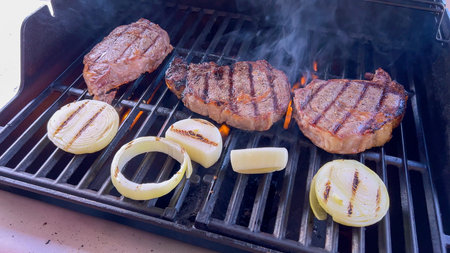 Ribeye steaks are sizzling alongside golden grilled onions on a barbecue grill, with wisps of smoke...