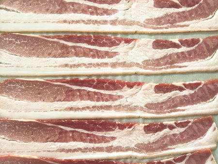 Neatly arranged raw bacon strips on a baking tray, prepared for cooking, capturing the fresh,...