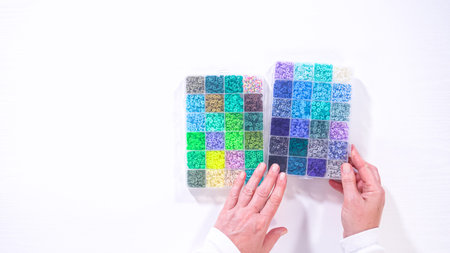 Flat lay. Woman hands gracefully poised over a collection of beads, sorted by color in transparent...