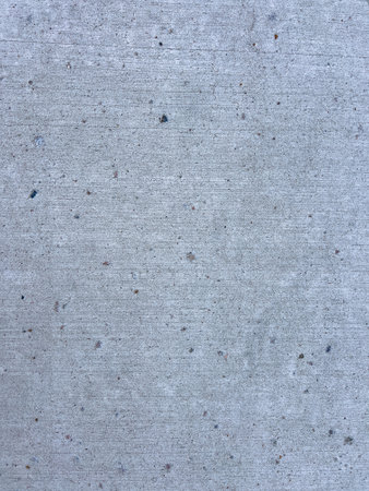 A detailed close-up of a concrete sidewalk surface, showcasing its fine texture and subtle imperfections. The concrete is light gray in color, with visible small stones and particles embedded within it. The surface displays faint vertical lines, indicating the brushing technique used during its finishing process. This image highlights the uniform yet varied texture of the concrete, capturing the nuances of its composition.の写真素材